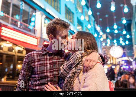 Romantic young couple kissing while standing against Christmas lights in city at night Stock Photo