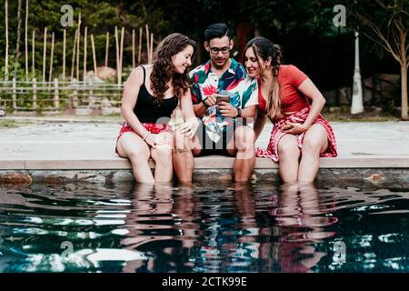 Friends using mobile phone while sitting at poolside Stock Photo