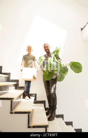 Smiling man with calathea and son carrying box while standing on staircase in new home Stock Photo