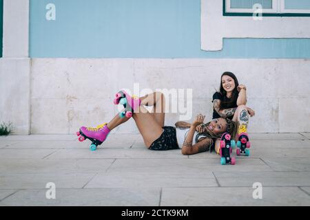 Smiling female friends with roller skates relaxing on footpath against wall in city Stock Photo
