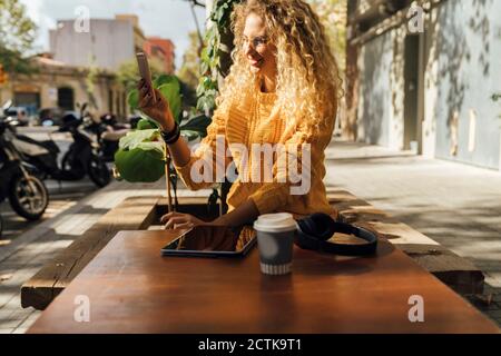 Smiling young female student photographing while sitting at sidewalk cafe in city Stock Photo