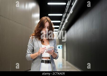 Redhead female entrepreneur using smart phone while standing in illuminated corridor Stock Photo