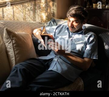 A relaxed man reading a book lying on a sofa by the window. Young latin man with black hair and a light blue shirt enjoying. Home alone. Stock Photo