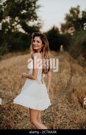 Smiling beautiful woman wearing white dress standing in wheat field during sunset Stock Photo