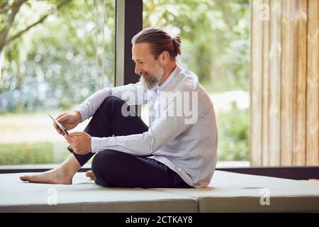 Smiling bearded mature man using digital tablet while sitting on bed by window at home Stock Photo