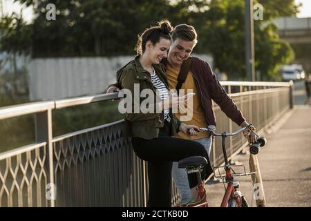 Smiling young couple looking at smart phone while standing with bicycle in city Stock Photo