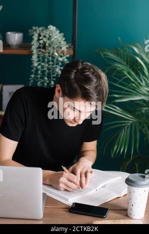 Young male student writing in book while sitting with laptop doing homework at table Stock Photo