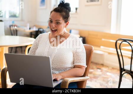 Shocked young woman with mouth open using laptop while sitting on chair in cafe Stock Photo