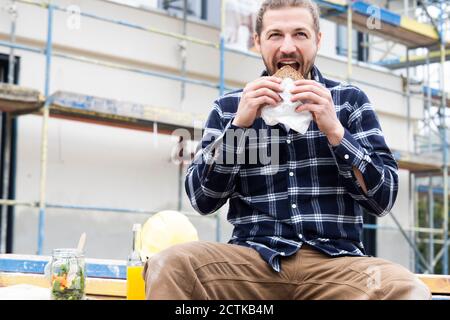 Construction worker eating bread while sitting against building at construction site Stock Photo