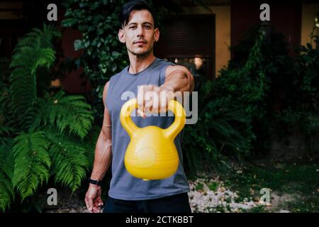 Mid adult man lifting kettlebell while standing against plants in yard Stock Photo