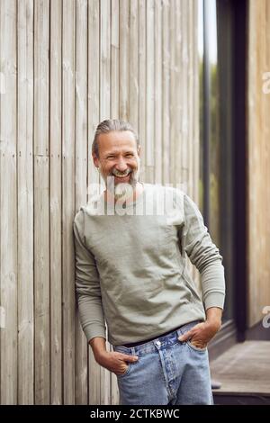 Happy bearded mature man with hands in pockets standing by wall at yard Stock Photo
