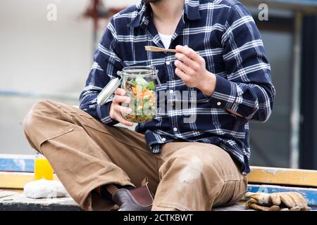 Close-up of construction worker holding salad in jar while sitting outdoors at site Stock Photo