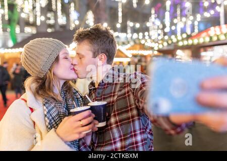 Couple holding hot chocolates kissing while taking selfie in Christmas market at night Stock Photo