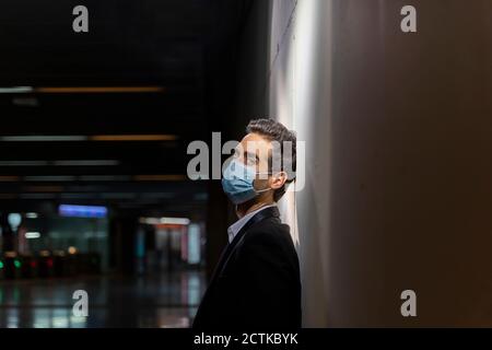 Male entrepreneur wearing mask while leaning on white wall at station Stock Photo