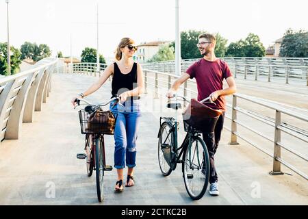 Happy couple talking while walking with bicycles on bridge against clear sky Stock Photo