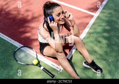 Smiling female tennis player listening music while sitting on floor in court Stock Photo