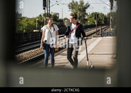 Smiling male and female friends talking while walking on railroad station platform Stock Photo