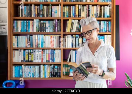 Active senior woman reading book while standing against bookshelf at home Stock Photo