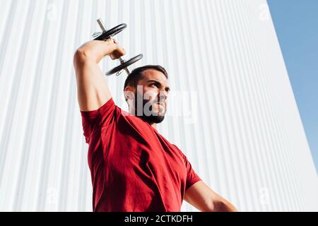 Young bearded man lifting dumbbell up while standing against wall Stock Photo