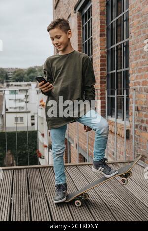 Boy using smart phone while standing with skateboard on boardwalk against railing Stock Photo