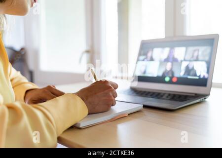 Cropped hands of female entrepreneur writing on diary while talking with colleagues through video conference at home Stock Photo