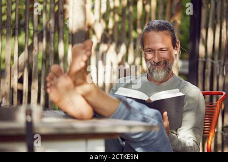 Smiling bearded man reading book while relaxing on chair in yard Stock Photo