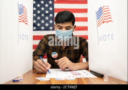 Young man in medical mask busy inside the polling booth with US flag as background - Concept of in person voting with covid-19 safety measure at US Stock Photo