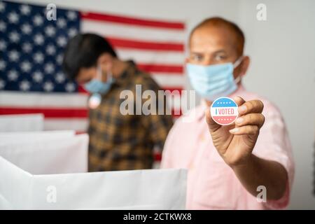 Man in medical mask showing I voted Sticker at polling booth with US flag as background - concept in person voting at US election. Stock Photo