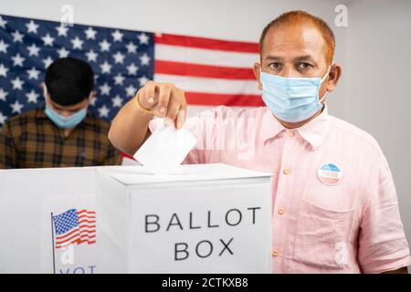 Man in medical mask placing ballot paper inside the ballot box while looking at camera - Concept of in person voting and people busy at polling booth Stock Photo