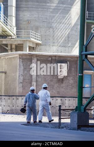 Two workers in white helmets and white protective suits with tools. On grey concrete factory building wall Stock Photo