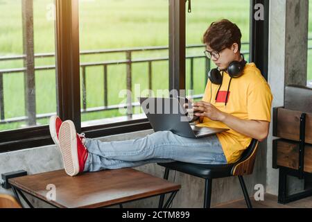 Asian freelance man chatting online on mobile phone and rewriting content in social network via laptop computer while resting in modern coffee shop, O Stock Photo