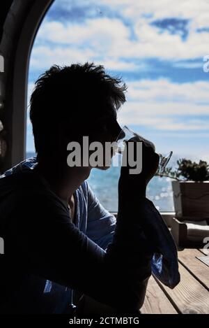 Silhouette of a young man finishing a cappuccino against the background of a window with a sea view. Stock Photo
