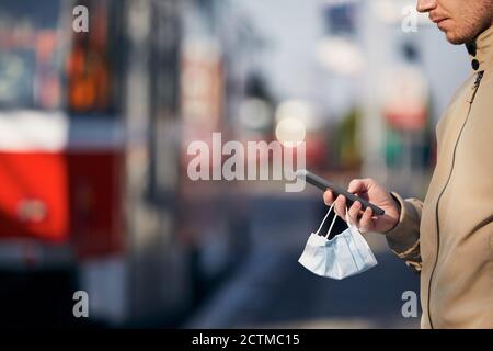 Young man with face mask in hand using smart phone during waiting for tram. Themes modern technology in new normal, coronavirus and personal protectio Stock Photo
