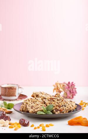 Homemade granola from oat flakes, dates, dried apricots, raisins, nuts in blue ceramic plate with a cup of coffee on a white and pink background. Side Stock Photo