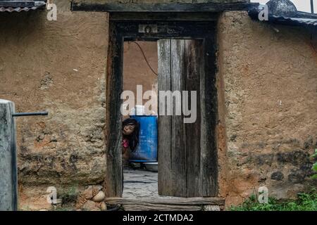 Bhutanese little girl peeking through the door of a traditional farmhouse in Punakha, Bhutan Stock Photo