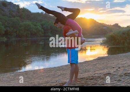 Fit athletic couple practicing acrobatics yoga at sunrise on a sandy beach with reflection of the sun on the calm water in a health and fitness or act Stock Photo