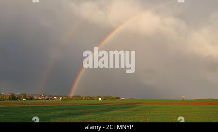 Strong double colored rainbow after thunderstorm with green fields and village in background near Plieningen, Stuttgart, Germany. Stock Photo