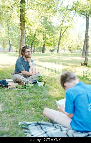 Boring young bearded teacher keeping pencil on mouth and waiting when students perform task in park Stock Photo