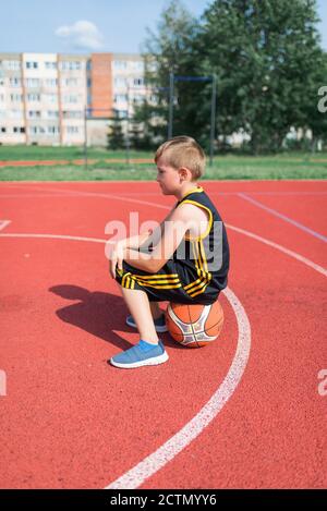 Side view of Sad elementary school boy sitting on basketball