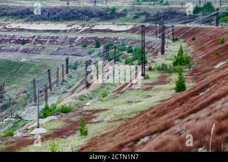Giant iron ore quarry in Kazakhstan.  Mining raw minerals for metal production. Abandoned part of quarry with electric masts, grass and fir trees. Stock Photo