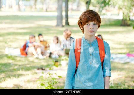 Portrait of serious redhead boy with satchel standing against classmates in public park Stock Photo