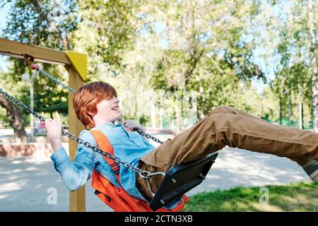 Excited carefree redhead boy with orange satchel swinging on swing in park after school Stock Photo