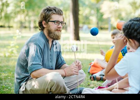 Young school teacher in eyeglasses sitting with crossed legs on grass and listening to pupils answers at outdoor astronomy class Stock Photo
