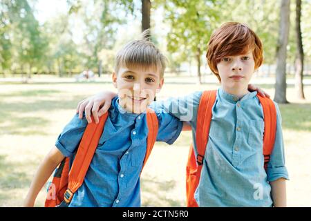 Portrait of serious and smiling school friends wearing orange satchels embracing each other in park Stock Photo