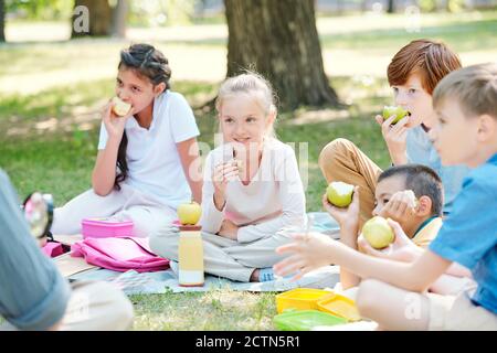 Group of positive kids sitting on plaid and eating apples while listening to teacher at outdoor lesson Stock Photo