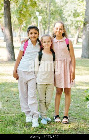 Three positive schoolgirls standing with satchels in park and embracing together Stock Photo