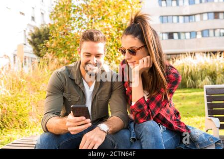 A happy couple using a application on their phone while sitting during summer on a park bench in the city. Stock Photo