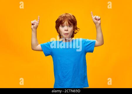 Red-haired boy grimaces face shows two hands up blue t-shirt yellow background  Stock Photo