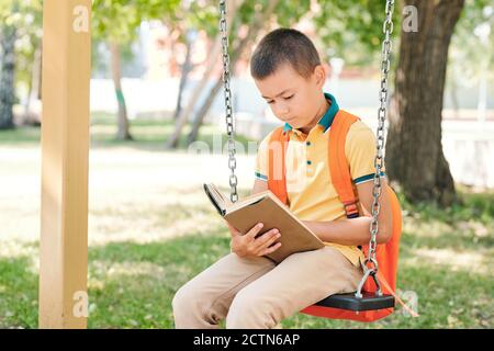 Curious boy with satchel on back sitting on swing in public park and reading literature while preparing for lesson Stock Photo
