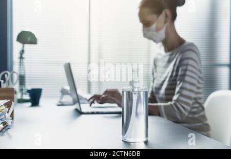 Woman sitting at office desk wearing a face mask and working with her laptop, hand sanitizer dispenser in the foreground, prevention and hygiene conce Stock Photo